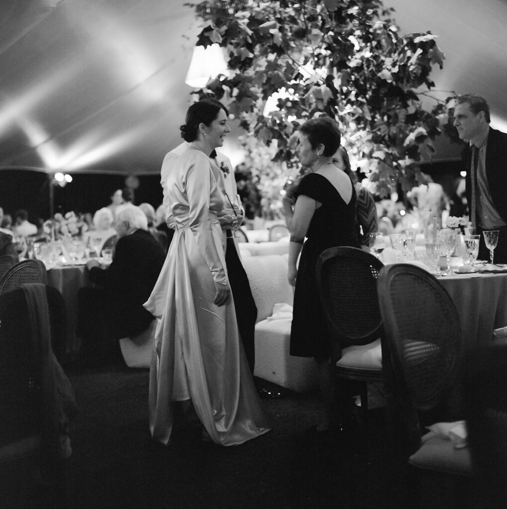 A mix of lush, garden-like floral centerpieces create a colorful display across tables under a canvas tent for a Sewanee wedding reception. Compotes, bud vases and ikebana arrangements overflow with garden roses, moab roses, icelandic poppies, lisianthus, tulips, dahlias, ranunculus, scabiosa, zinnias, sweet peas, oncidium and more in shades of peach, red, apricot and yellow. Lush greenery is suspended around light pendants under the tent. Design by Rosemary and Finch Floral Design in Nashville, TN.
