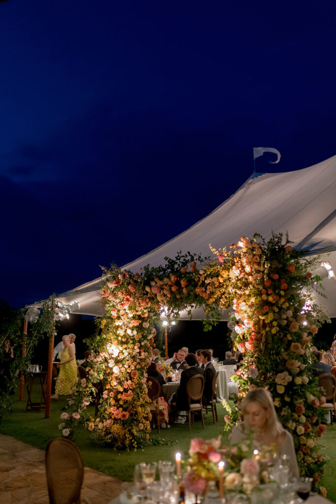 A lush, colorful floral arch frames an elegant tented wedding in Sewanee, TN; Overflowing with garden roses, spray roses, ranunculus, sweet peas, moab roses, lisianthus, yarrow, zinnias, tulips, butterfly ranunculus, foxglove, smokebush, oncidium and greenery in soft shades of blush, peach, butter-yellow and apricot. The floral arch curates a romantic, garden-inspired atmosphere with natural, airy textures and timeless charm. Design by Rosemary and Finch Floral Design in Nashville, TN.