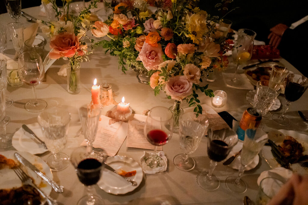 A mix of lush, garden-like floral centerpieces create a colorful display across tables under a canvas tent for a Sewanee wedding reception. Compotes, bud vases and ikebana arrangements overflow with garden roses, moab roses, icelandic poppies, lisianthus, tulips, dahlias, ranunculus, scabiosa, zinnias, sweet peas, oncidium and more in shades of peach, red, apricot and yellow. Lush greenery is suspended around light pendants under the tent. Design by Rosemary and Finch Floral Design in Nashville, TN.
