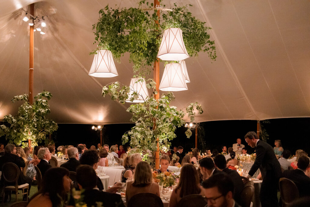 A mix of lush, garden-like floral centerpieces create a colorful display across tables under a canvas tent for a Sewanee wedding reception. Compotes, bud vases and ikebana arrangements overflow with garden roses, moab roses, icelandic poppies, lisianthus, tulips, dahlias, ranunculus, scabiosa, zinnias, sweet peas, oncidium and more in shades of peach, red, apricot and yellow. Lush greenery is suspended around light pendants under the tent. Design by Rosemary and Finch Floral Design in Nashville, TN.
