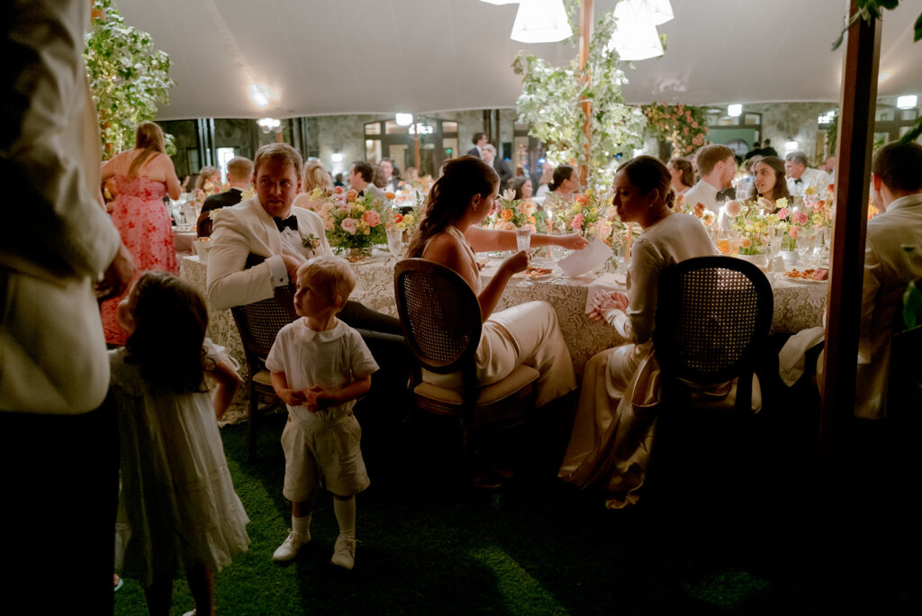 A mix of lush, garden-like floral centerpieces create a colorful display across tables under a canvas tent for a Sewanee wedding reception. Compotes, bud vases and ikebana arrangements overflow with garden roses, moab roses, icelandic poppies, lisianthus, tulips, dahlias, ranunculus, scabiosa, zinnias, sweet peas, oncidium and more in shades of peach, red, apricot and yellow. Lush greenery is suspended around light pendants under the tent. Design by Rosemary and Finch Floral Design in Nashville, TN.
