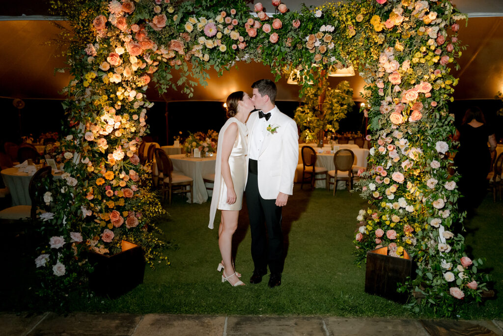 A lush, colorful floral arch frames an elegant tented wedding in Sewanee, TN; Overflowing with garden roses, spray roses, ranunculus, sweet peas, moab roses, lisianthus, yarrow, zinnias, tulips, butterfly ranunculus, foxglove, smokebush, oncidium and greenery in soft shades of blush, peach, butter-yellow and apricot. The floral arch curates a romantic, garden-inspired atmosphere with natural, airy textures and timeless charm. Design by Rosemary and Finch Floral Design in Nashville, TN.