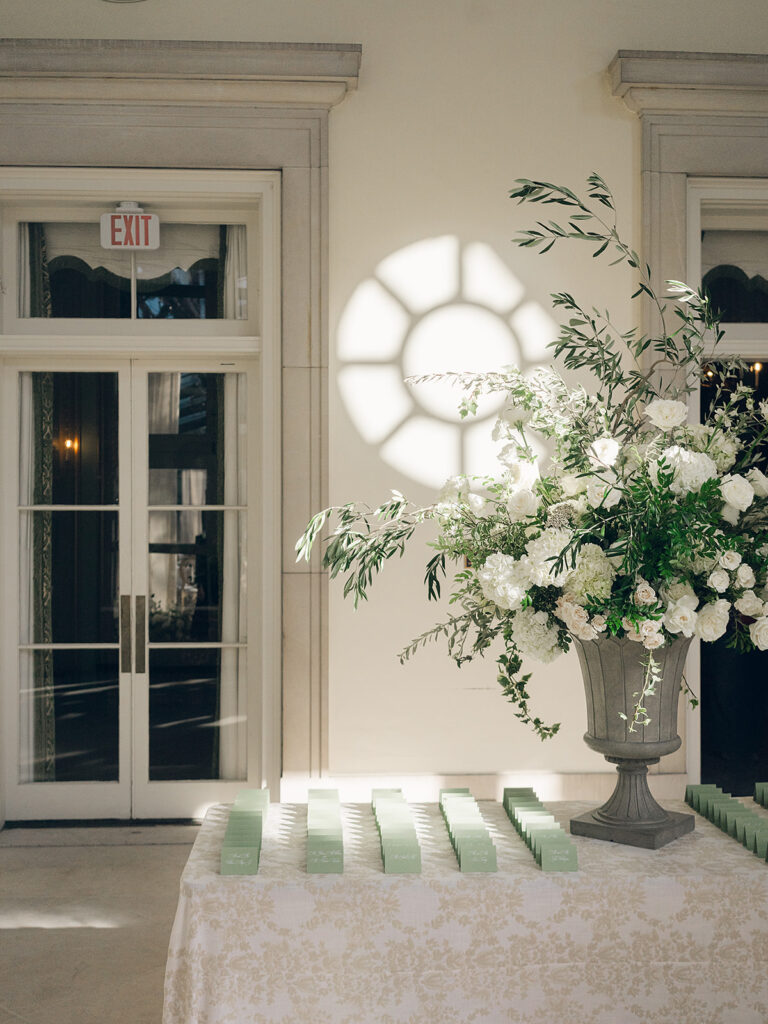 Soft, elegant floral statements create a stunning focal point for guests as they enter a glass-tented reception. All-white hydrangeas, lisianthus, cloud roses, spray roses, delphinium, stock, sweet peas, and dahlias spill over the sides of a loggia fountain. A large statement urn with all-white florals matching the fountain creates an extravagant moment for the escort display table. Design by Rosemary & Finch Floral Design in Nashville, TN.