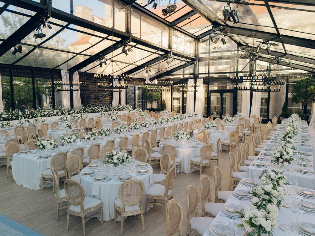 Romantic and stunning glass-tented wedding reception at Cheekwood botanical garden in Nashville, TN. Elegant garden-inspired centerpieces feature lush, all-white cloud roses, hydrangeas, lisianthus, dahlias, sweet peas, double tulips, delphinium, stock, allium, flowering champagne, and more with light greenery. Matching banquette meadows line either side of the glass tent creating a lush and timeless feel. Design by Rosemary & Finch Floral Design in Nashville, TN.