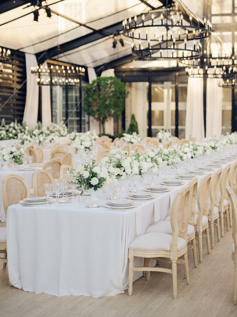 Romantic and stunning glass-tented wedding reception at Cheekwood botanical garden in Nashville, TN. Elegant garden-inspired centerpieces feature lush, all-white cloud roses, hydrangeas, lisianthus, dahlias, sweet peas, double tulips, delphinium, stock, allium, flowering champagne, and more with light greenery. Matching banquette meadows line either side of the glass tent creating a lush and timeless feel. Design by Rosemary & Finch Floral Design in Nashville, TN.
