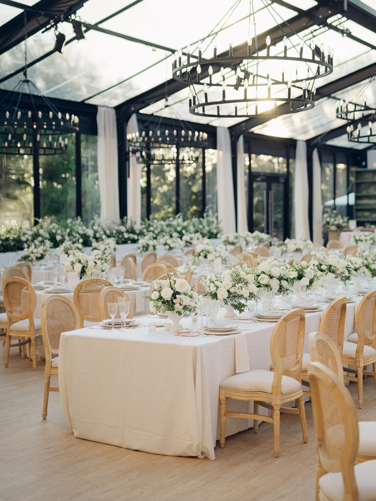 Romantic and stunning glass-tented wedding reception at Cheekwood botanical garden in Nashville, TN. Elegant garden-inspired centerpieces feature lush, all-white cloud roses, hydrangeas, lisianthus, dahlias, sweet peas, double tulips, delphinium, stock, allium, flowering champagne, and more with light greenery. Matching banquette meadows line either side of the glass tent creating a lush and timeless feel. Design by Rosemary & Finch Floral Design in Nashville, TN.