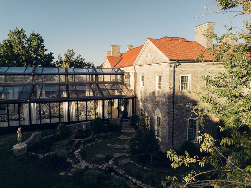 Romantic and stunning glass-tented wedding reception at Cheekwood botanical garden in Nashville, TN. Elegant garden-inspired centerpieces feature lush, all-white cloud roses, hydrangeas, lisianthus, dahlias, sweet peas, double tulips, delphinium, stock, allium, flowering champagne, and more with light greenery. Matching banquette meadows line either side of the glass tent creating a lush and timeless feel. Design by Rosemary & Finch Floral Design in Nashville, TN.