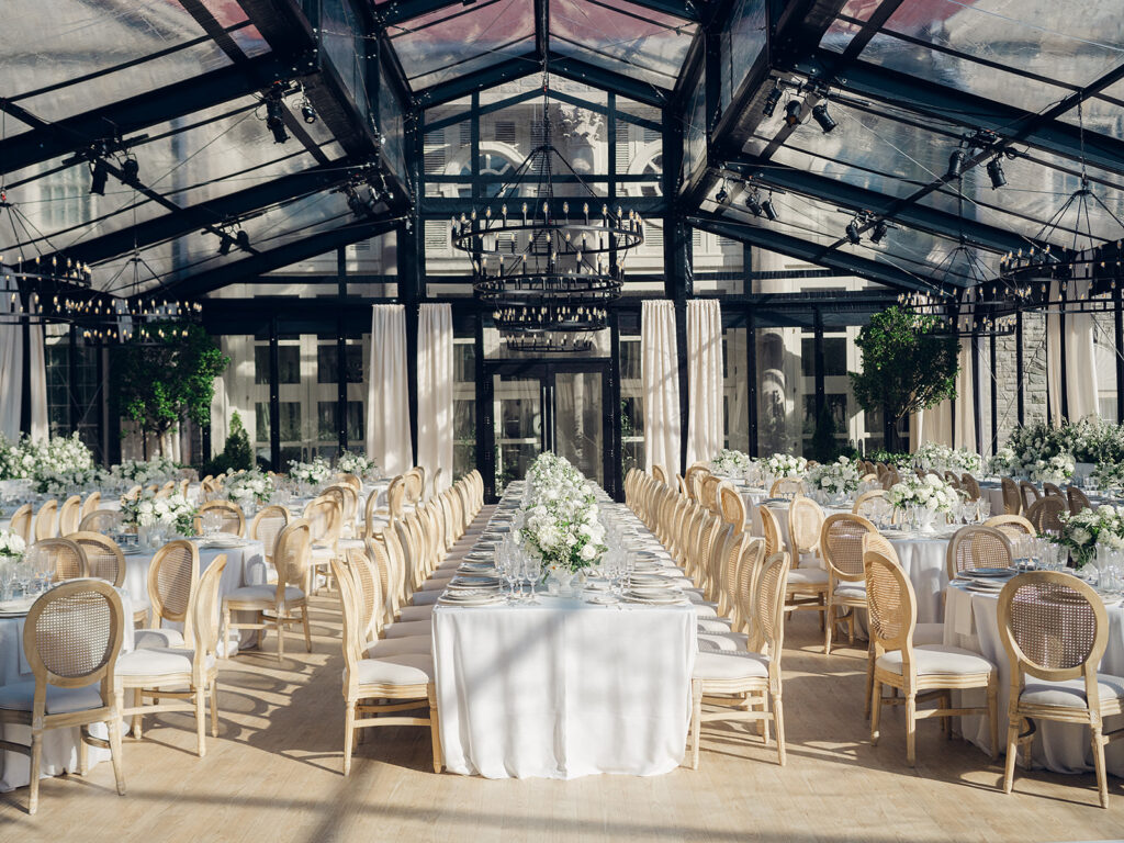 Romantic and stunning glass-tented wedding reception at Cheekwood botanical garden in Nashville, TN. Elegant garden-inspired centerpieces feature lush, all-white cloud roses, hydrangeas, lisianthus, dahlias, sweet peas, double tulips, delphinium, stock, allium, flowering champagne, and more with light greenery. Matching banquette meadows line either side of the glass tent creating a lush and timeless feel. Design by Rosemary & Finch Floral Design in Nashville, TN.