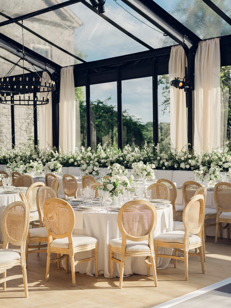 Romantic and stunning glass-tented wedding reception at Cheekwood botanical garden in Nashville, TN. Elegant garden-inspired centerpieces feature lush, all-white cloud roses, hydrangeas, lisianthus, dahlias, sweet peas, double tulips, delphinium, stock, allium, flowering champagne, and more with light greenery. Matching banquette meadows line either side of the glass tent creating a lush and timeless feel. Design by Rosemary & Finch Floral Design in Nashville, TN.