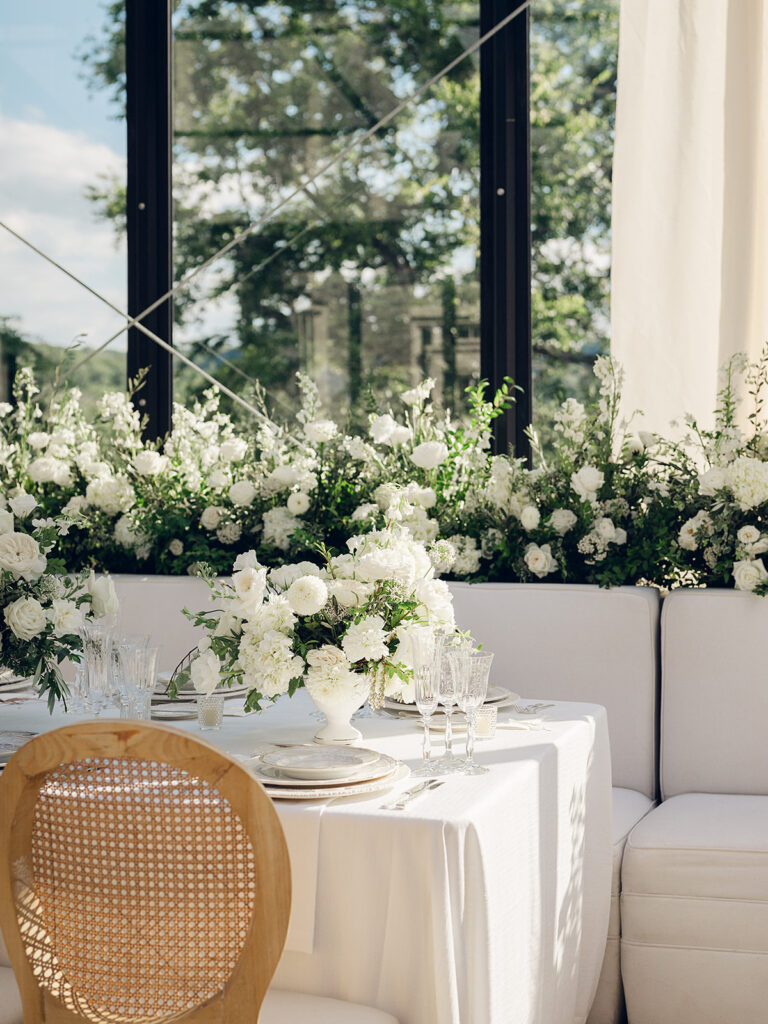 Romantic and stunning glass-tented wedding reception at Cheekwood botanical garden in Nashville, TN. Elegant garden-inspired centerpieces feature lush, all-white cloud roses, hydrangeas, lisianthus, dahlias, sweet peas, double tulips, delphinium, stock, allium, flowering champagne, and more with light greenery. Matching banquette meadows line either side of the glass tent creating a lush and timeless feel. Design by Rosemary & Finch Floral Design in Nashville, TN.