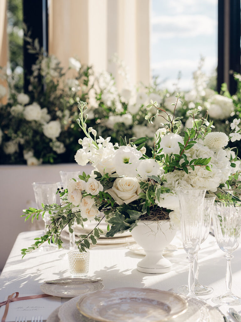 Romantic and stunning glass-tented wedding reception at Cheekwood botanical garden in Nashville, TN. Elegant garden-inspired centerpieces feature lush, all-white cloud roses, hydrangeas, lisianthus, dahlias, sweet peas, double tulips, delphinium, stock, allium, flowering champagne, and more with light greenery. Matching banquette meadows line either side of the glass tent creating a lush and timeless feel. Design by Rosemary & Finch Floral Design in Nashville, TN.