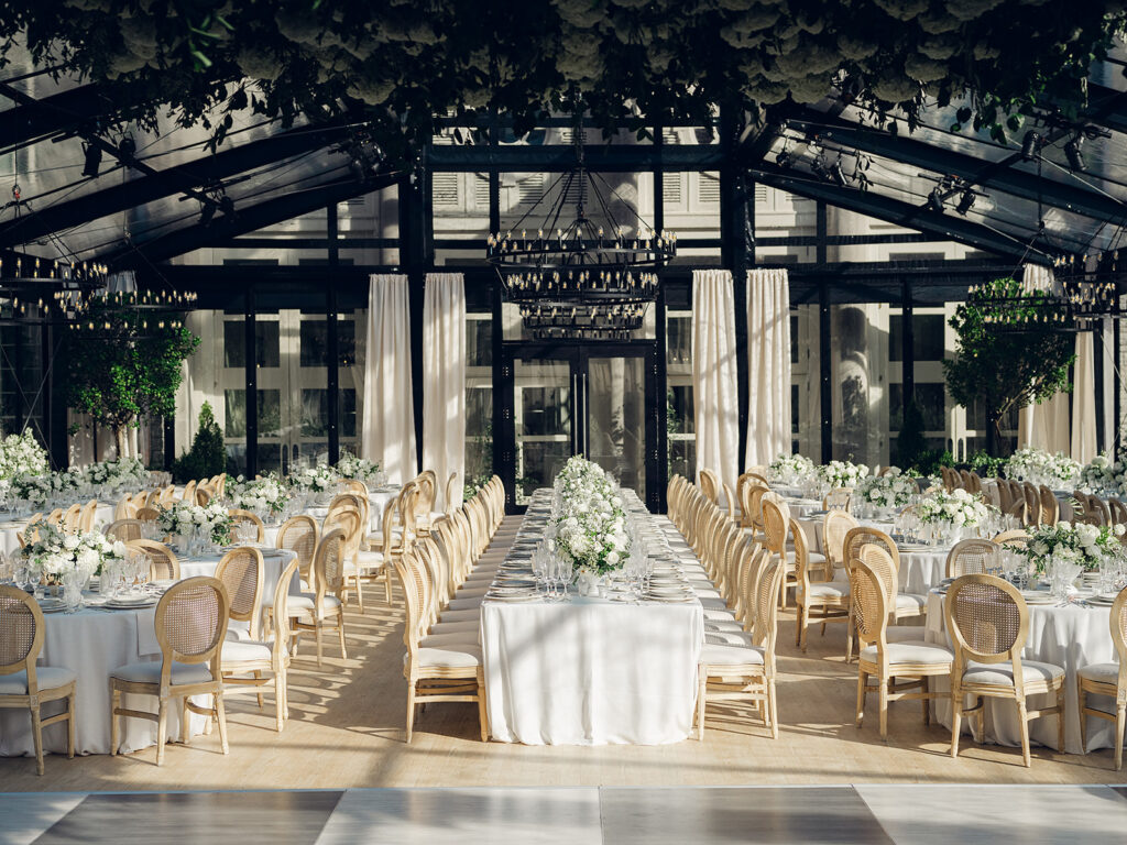 Romantic and stunning glass-tented wedding reception at Cheekwood botanical garden in Nashville, TN. Elegant garden-inspired centerpieces feature lush, all-white cloud roses, hydrangeas, lisianthus, dahlias, sweet peas, double tulips, delphinium, stock, allium, flowering champagne, and more with light greenery. Matching banquette meadows line either side of the glass tent creating a lush and timeless feel. Design by Rosemary & Finch Floral Design in Nashville, TN.