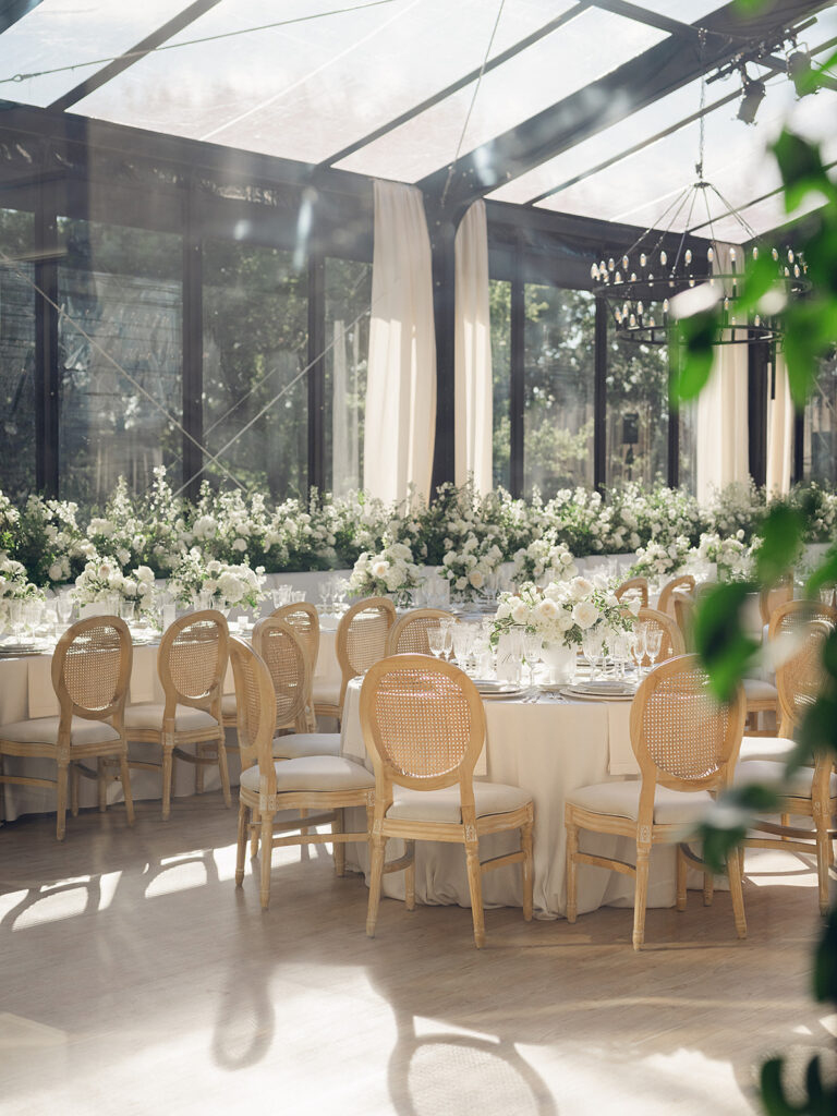 Romantic and stunning glass-tented wedding reception at Cheekwood botanical garden in Nashville, TN. Elegant garden-inspired centerpieces feature lush, all-white cloud roses, hydrangeas, lisianthus, dahlias, sweet peas, double tulips, delphinium, stock, allium, flowering champagne, and more with light greenery. Matching banquette meadows line either side of the glass tent creating a lush and timeless feel. Design by Rosemary & Finch Floral Design in Nashville, TN.