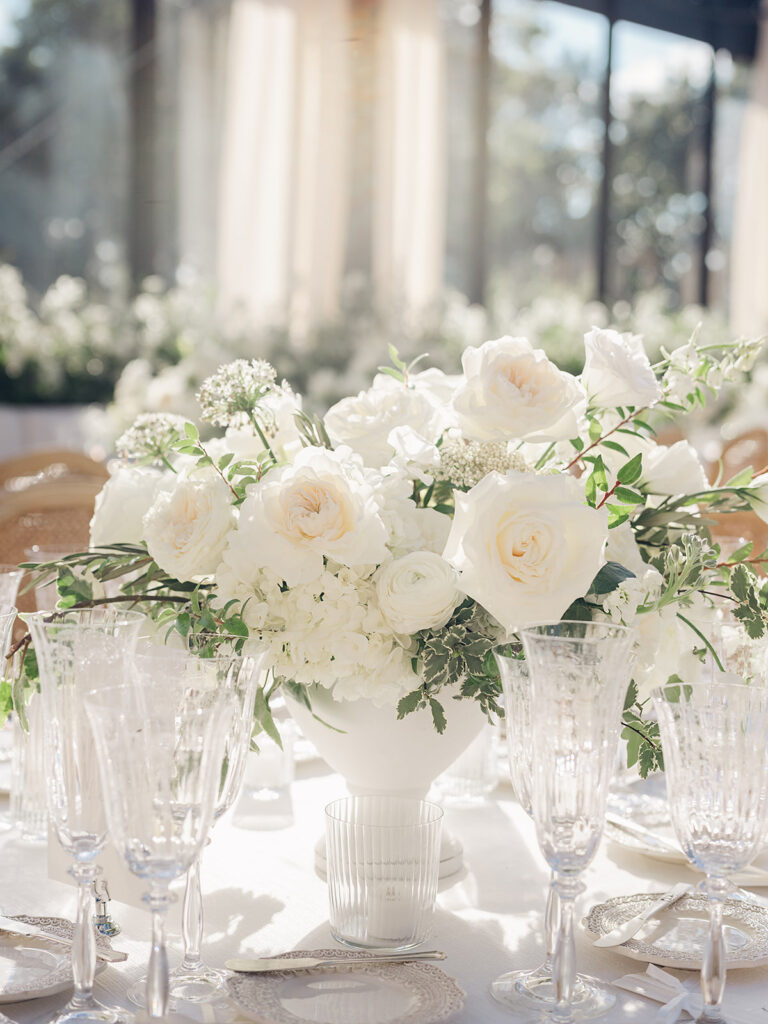 Romantic and stunning glass-tented wedding reception at Cheekwood botanical garden in Nashville, TN. Elegant garden-inspired centerpieces feature lush, all-white cloud roses, hydrangeas, lisianthus, dahlias, sweet peas, double tulips, delphinium, stock, allium, flowering champagne, and more with light greenery. Matching banquette meadows line either side of the glass tent creating a lush and timeless feel. Design by Rosemary & Finch Floral Design in Nashville, TN.