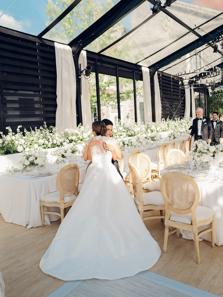 Romantic and stunning glass-tented wedding reception at Cheekwood botanical garden in Nashville, TN. Elegant garden-inspired centerpieces feature lush, all-white cloud roses, hydrangeas, lisianthus, dahlias, sweet peas, double tulips, delphinium, stock, allium, flowering champagne, and more with light greenery. Matching banquette meadows line either side of the glass tent creating a lush and timeless feel. Design by Rosemary & Finch Floral Design in Nashville, TN.